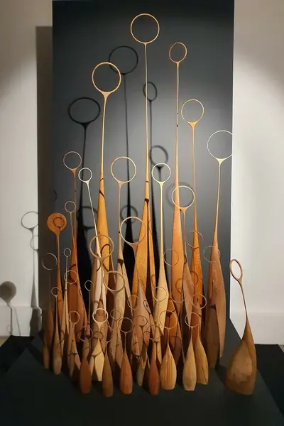 Bosque de piezas de madera torneadas con aros en lo alto, sobre una peana oscura en una galería