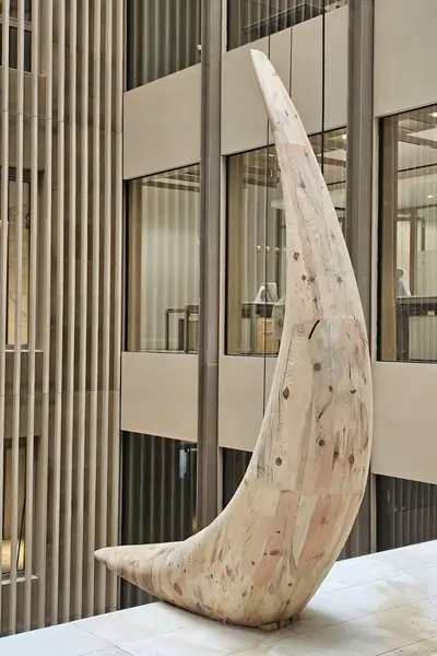 Escultura de madera clara con forma de media luna, expuesta ante una fachada de columnas blancas