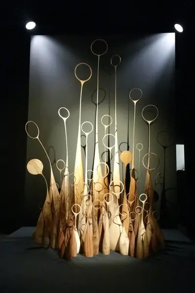 Instalación de esculturas de madera con aros metálicos en lo alto, iluminada sobre fondo oscuro