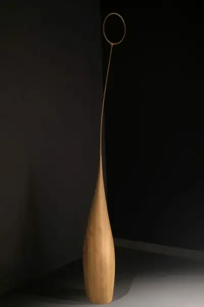 Escultura alta de madera clara, con un aro en lo alto, sobre fondo negro