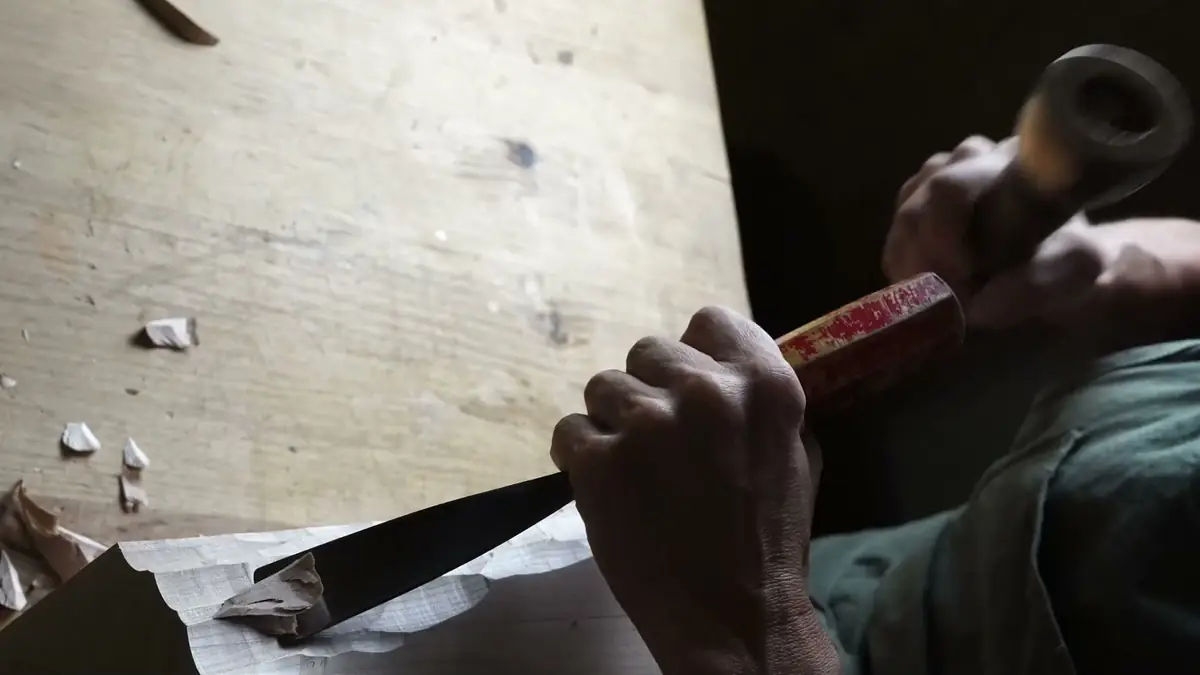 Las manos de Sandra Krysiak empujando la gubia con el mazo sobre un bloque de madera, entre virutas, en el banco del taller