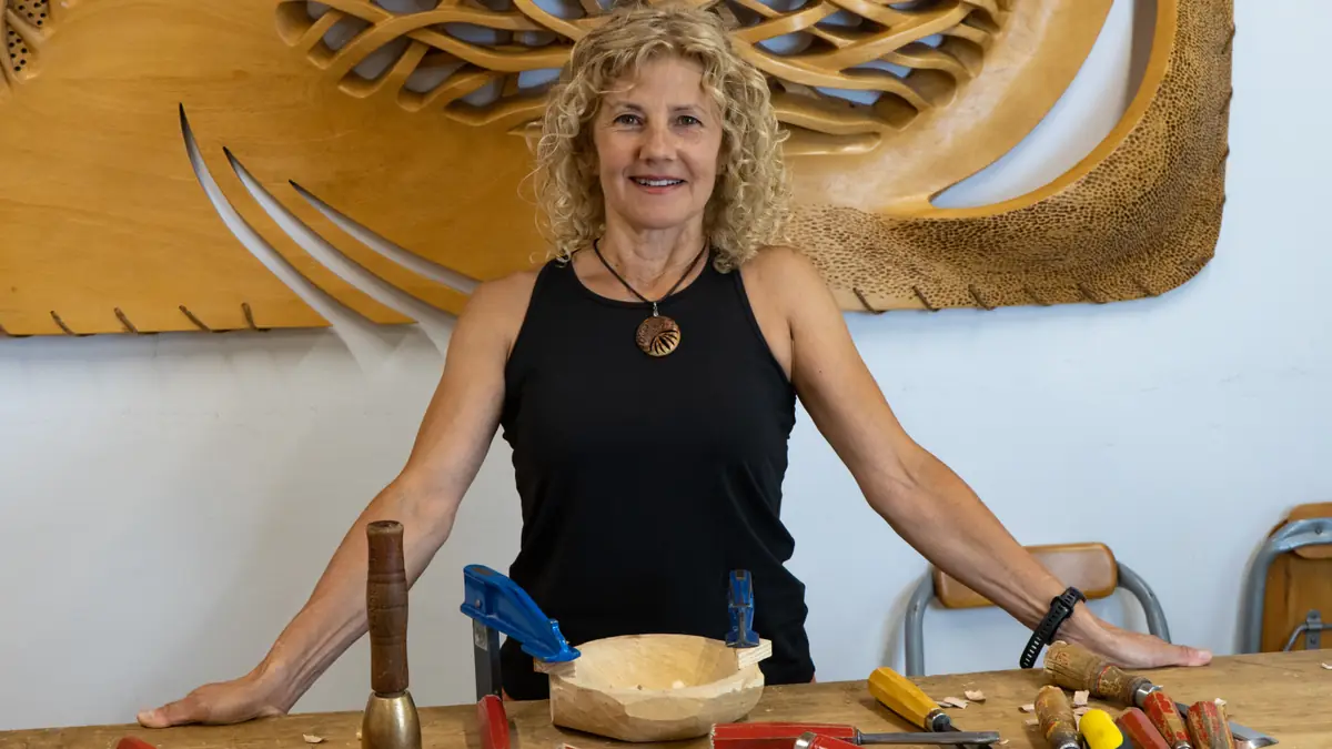 Sandra Krysiak tallando un cuenco con gubia y mazo en su taller, con un relieve de madera al fondo