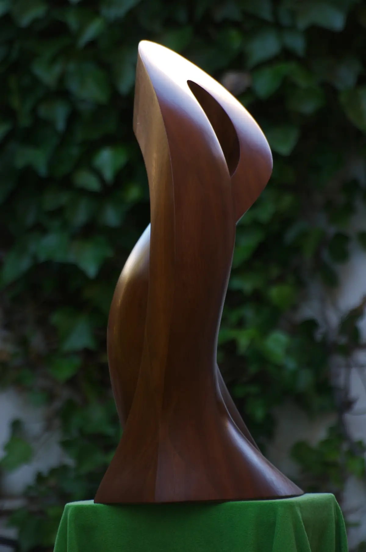Escultura vertical de iroko con una forma curva y una abertura cerca de la parte superior, fotografiada ante un jardín