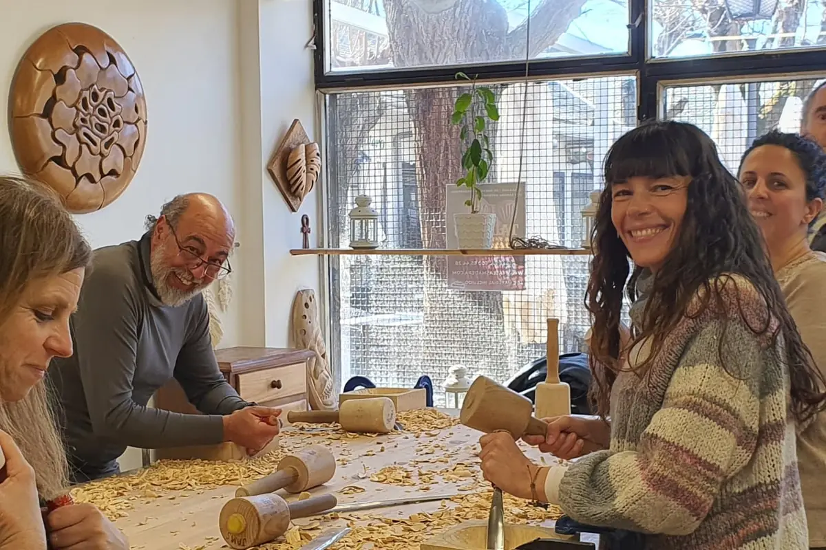 Grupo de alumnos tallando con mazo y gubia alrededor de la mesa del taller, cubierta de virutas, junto al ventanal