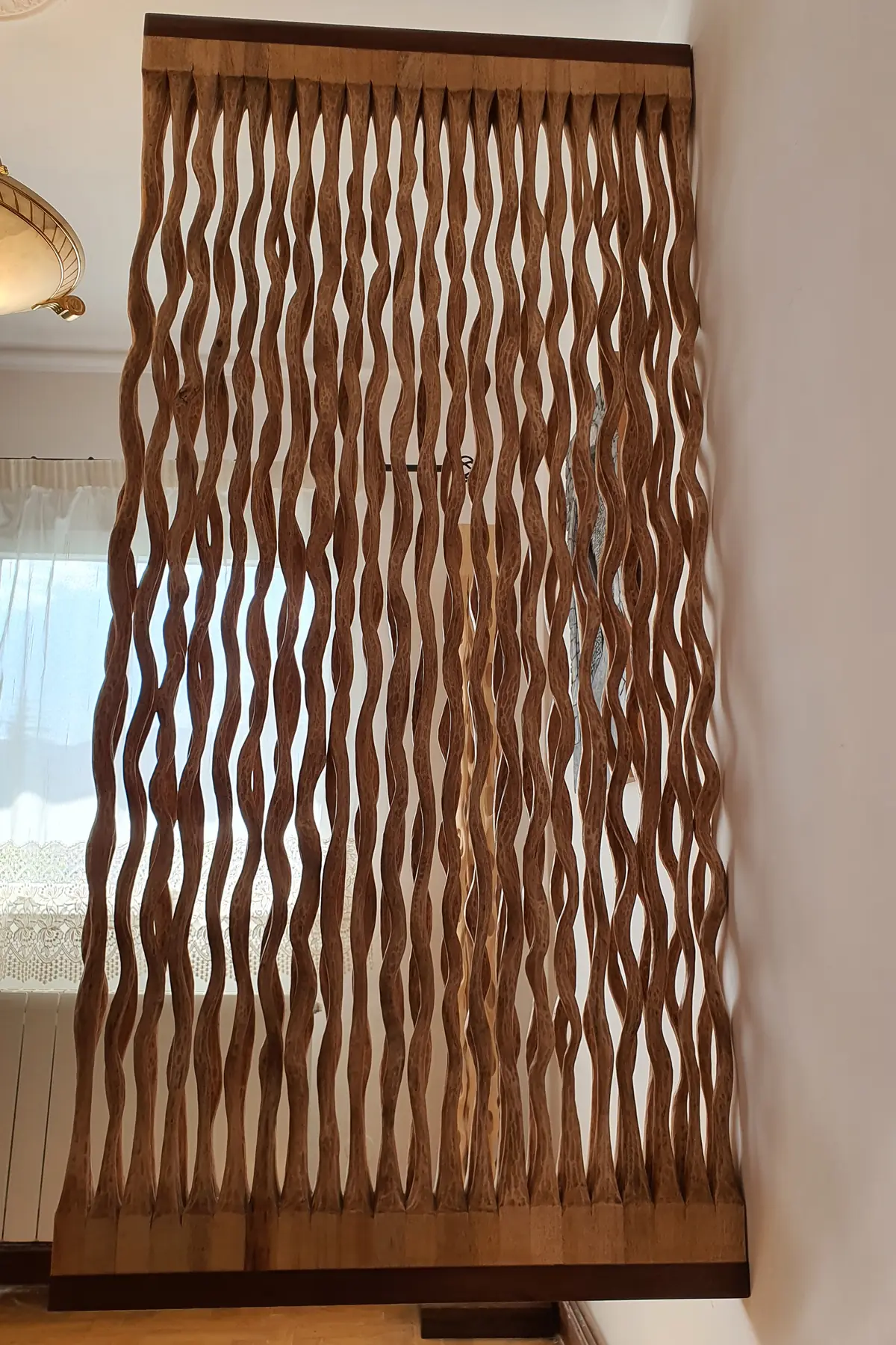 Panel vertical de madera de ayous formado por listones ondulados tallados uno a uno, colgado en una pared junto a una ventana