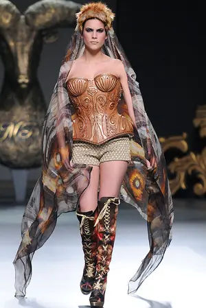 Una modelo desfila en la pasarela con el corsé de madera tallada y un tocado de pelo, bajo una cabeza de carnero dorada