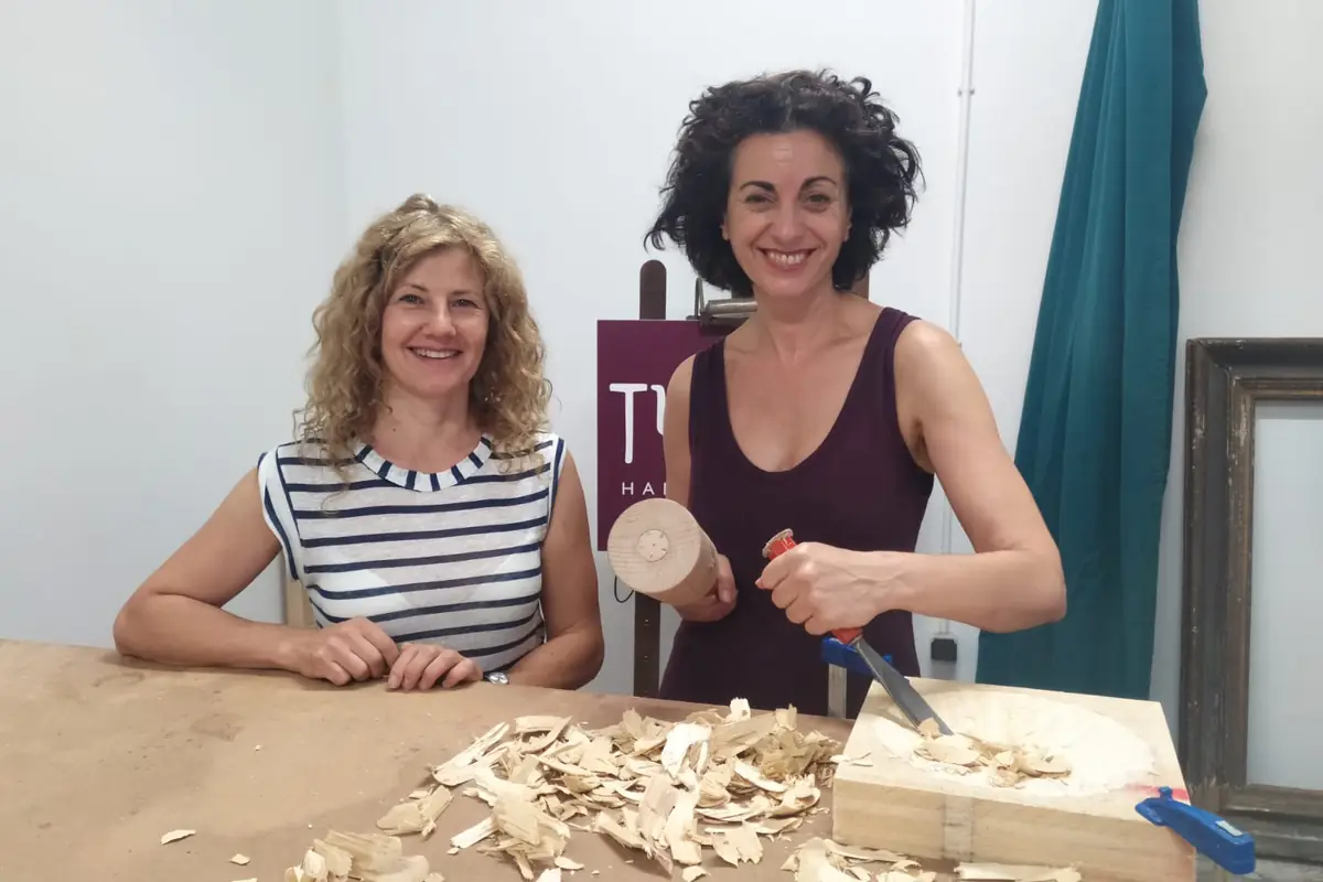 Sandra Krysiak y la actriz Paloma Vidal en una mesa cubierta de virutas, con las gubias y una pieza de madera recién tallada